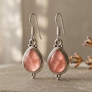 Puede incluir: Un par de pendientes colgantes de plata con piedras rosas translúcidas en forma de lágrima engastadas en marcos de metal pulido. Cada pendiente cuelga de un gancho de plata sencillo e incluye detalles de cuentas metálicas en la base.