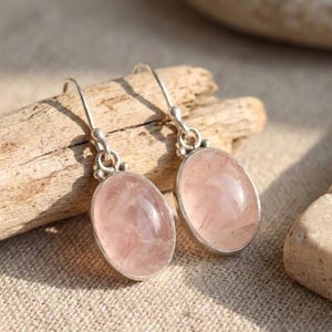 May include: A pair of drop earrings featuring oval-shaped, translucent pale pink rose quartz gemstones set in polished silver-toned metal frames. Each earring hangs from a simple silver-toned hook, showcasing a smooth, rounded cabochon cut.