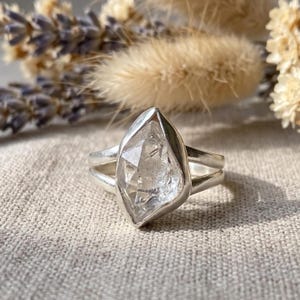 Puede incluir: Un anillo de plata con una piedra preciosa transparente, facetada y en forma de marquesa, engastada en un bisel de metal liso. La banda está formada por dos hilos de plata paralelos que se unen en el engaste, creando un diseño de brazo dividido.