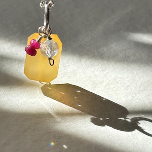 May include: A silver chain necklace featuring a rectangular yellow gemstone pendant. Attached to the silver bail are two small, round, faceted deep pink rubies and one clear, faceted, round crystal bead, all suspended together on the silver chain.