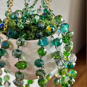 May include: A hanging planter featuring a small white ceramic pot filled with green moss and cascading strands of faceted green and clear crystal beads. The plant is suspended by gold-toned metal chains attached to the rim of the pot.