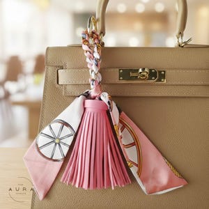 May include: A tan leather handbag with a gold clasp and handle. A pink leather tassel and a patterned scarf are attached to the handle. The scarf has a white background with a wheel design and gold trim.