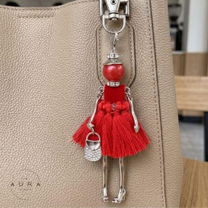 Pode incluir: Um chaveiro vermelho em forma de boneca com braços, pernas e saltos altos prateados. A boneca tem um vestido vermelho com uma saia de borlas e uma pequena bolsa prateada. O chaveiro está preso a uma bolsa de couro bege.