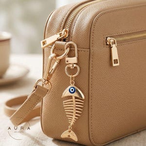 May include: A gold-colored fishbone charm with a blue eye detail hangs from a tan handbag. The charm is attached to the bag's zipper with a gold clasp. The handbag has a textured surface and gold zippers.