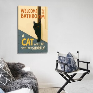 May include: A canvas print with the text "WELCOME TO THE BATHROOM" and "A CAT WILL BE WITH YOU SHORTLY". A black cat with yellow eyes peeks out from behind a door. The background is a light yellow with a blue and gray floor.