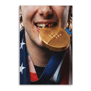 May include: Close-up of a person smiling, biting a gold medal. The medal features the Olympic rings and is on a blue and white ribbon. The person is wearing a black shirt and a draped American flag.