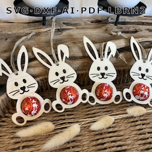 Può includere: Quattro decorazioni pasquali a forma di coniglietto in legno, ognuna con un uovo di cioccolato avvolto in carta rossa. I coniglietti hanno dettagli neri e sono appesi a un filo. Su un cestino intrecciato con fiori secchi.