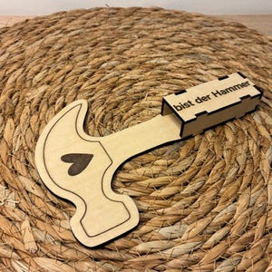 Op de afbeelding: Een houten hamer-vormige doos met een hart uitsparing en de tekst "bist der Hammer" op de doos. De hamer is lichtbruin met een donkerbruin hart. De doos ligt op een geweven, cirkelvormig, bruin oppervlak.