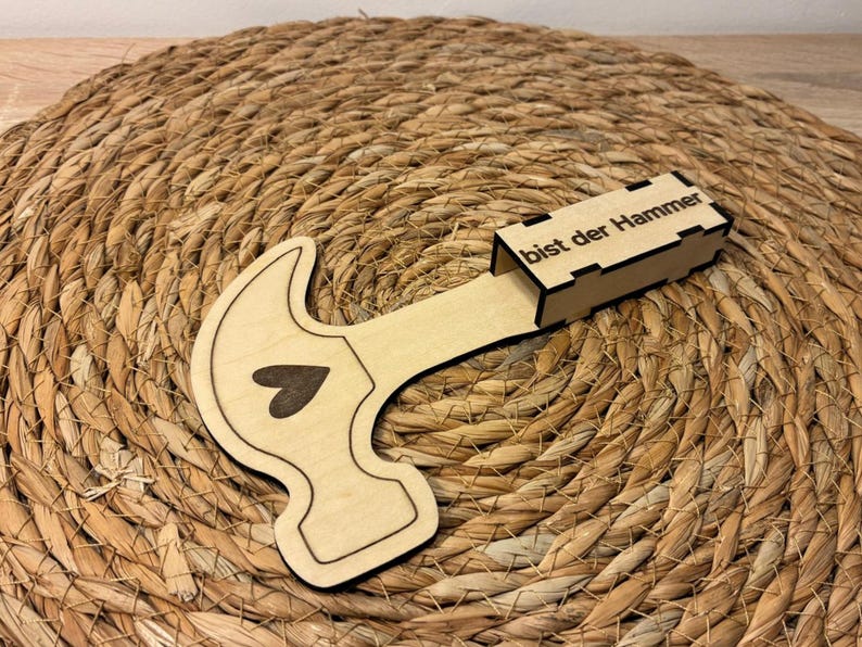 Op de afbeelding: Een houten hamer-vormige doos met een hart uitsparing en de tekst "bist der Hammer" op de doos. De hamer is lichtbruin met een donkerbruin hart. De doos ligt op een geweven, cirkelvormig, bruin oppervlak.