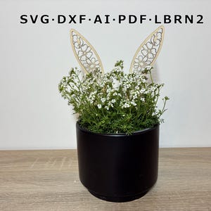 Puede incluir: Una maceta cilíndrica negra llena de vegetación y flores blancas, adornada con dos orejas de conejo de madera. Las orejas están cortadas con láser con un diseño floral. El texto en la parte superior dice: SVG•DXF•AI•PDF•LBRN2.