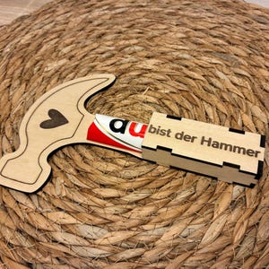Op de afbeelding: Een houten hamer-vormige doos met een hart uitsparing en de tekst "a bist der Hammer" op een rechthoekige doos. De hamer bevat een tube met rood en wit product. De items liggen op een geweven, cirkelvormig oppervlak.
