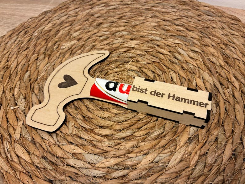 Op de afbeelding: Een houten hamer-vormige doos met een hart uitsparing en de tekst "a bist der Hammer" op een rechthoekige doos. De hamer bevat een tube met rood en wit product. De items liggen op een geweven, cirkelvormig oppervlak.