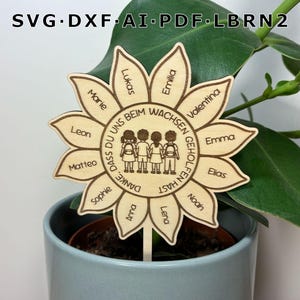 Può includere: Un segnaposto per piante in legno chiaro a forma di girasole con il testo inciso 'DANKE, DASS DU UNS BEIM WACHSEN GEHOLFEN HAST' che circonda un'illustrazione centrale di quattro bambini, con nomi individuali su ogni petalo.