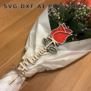 Könnte beinhalten: Ein Strauß aus orangefarbenen Rosen und weißem Schleierkraut, eingewickelt in weißes Papier. Eine Holzrose mit dem Wort "Mama" ist am Strauß befestigt. Die Holzrose ist rot und grün.