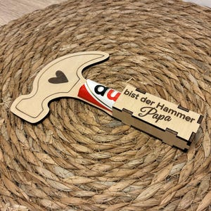 Op de afbeelding: Een houten cadeauverpakking in de vorm van een hamer met een hartdetail en de tekst "bist der Hammer Papa". De hamer bevat een rood-witte chocoladereep. De items liggen op een geweven, cirkelvormig oppervlak.