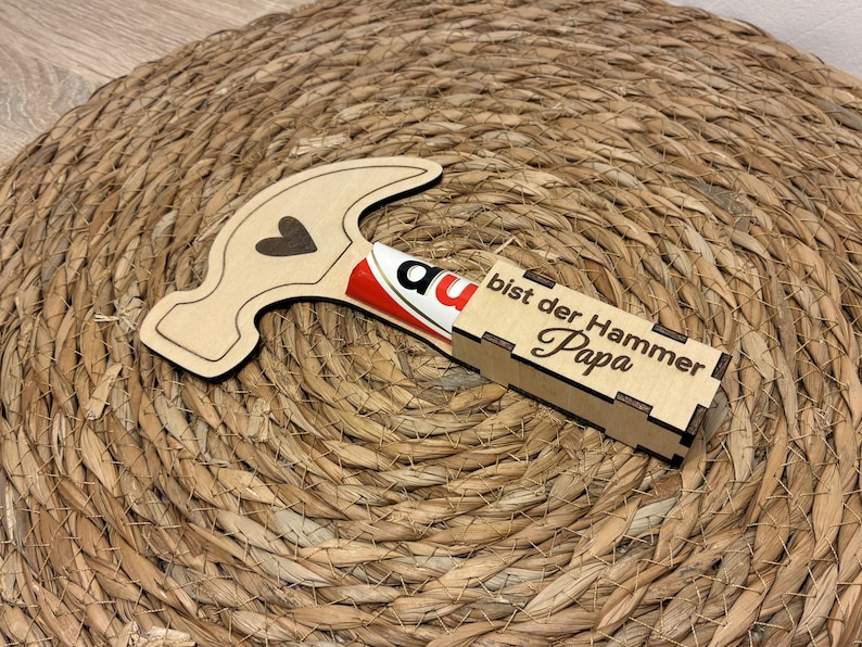 Op de afbeelding: Een houten cadeauverpakking in de vorm van een hamer met een hartdetail en de tekst "bist der Hammer Papa". De hamer bevat een rood-witte chocoladereep. De items liggen op een geweven, cirkelvormig oppervlak.