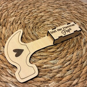 Op de afbeelding: Een houten hamer-vormige doos met een hart ontwerp en de tekst "bist der Hammer Papa". De doos is gemaakt van lichtgekleurd hout en ligt op een geweven, bruin oppervlak.