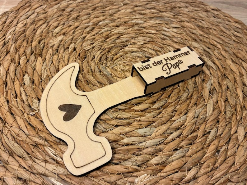 Op de afbeelding: Een houten hamer-vormige doos met een hart ontwerp en de tekst "bist der Hammer Papa". De doos is gemaakt van lichtgekleurd hout en ligt op een geweven, bruin oppervlak.