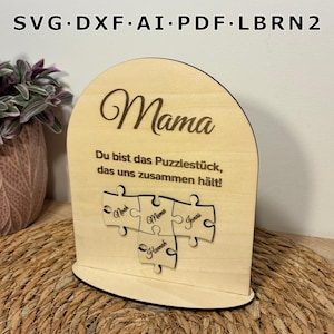 Könnte beinhalten: Ein Holzschild in Form eines Puzzleteils mit dem Wort "Mama" in Schreibschrift. Das Schild enthält den Text "Du bist das Puzzlestück, das uns zusammen hält!" und drei Puzzleteile mit Namen.