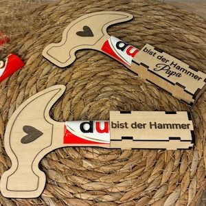 Op de afbeelding: Twee houten hamervormige cadeaus met een hartontwerp, elk met een chocoladereep. De tekst "bist der Hammer Papa" is met laser uitgesneden op de geschenkdoos. De achtergrond is een geweven, natuurlijk gekleurde mat.