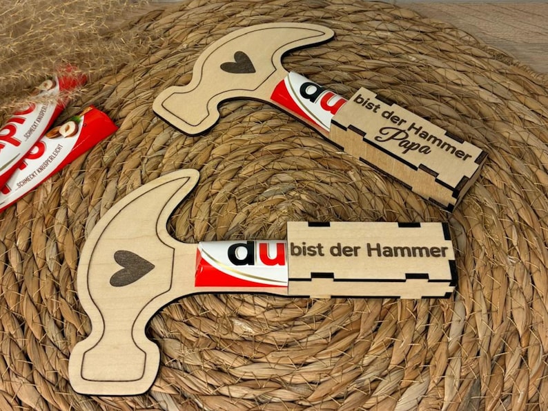 Op de afbeelding: Twee houten hamervormige cadeaus met een hartontwerp, elk met een chocoladereep. De tekst "bist der Hammer Papa" is met laser uitgesneden op de geschenkdoos. De achtergrond is een geweven, natuurlijk gekleurde mat.