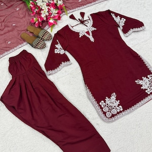 May include: A maroon salwar kameez set featuring a tunic with white floral embroidery on the neckline, sleeves, and hem, paired with matching pleated trousers, a sheer maroon dupatta with white border detailing, and two embroidered flat shoes.