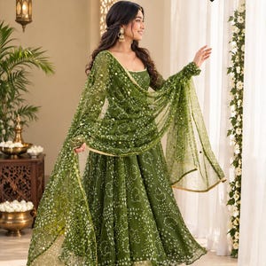 May include: An olive green floor-length dress featuring a white bandhani tie-dye pattern and gold trim. The outfit includes a matching sheer dupatta with gold sequin embellishments. The text SharvilStyles is visible in the upper right corner.