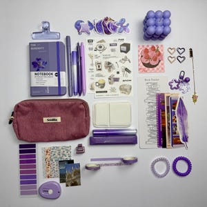 Könnte beinhalten: Eine Auswahl an lila und malvenfarbenen Schreibwaren, darunter ein Notizbuch, Stifte, Aufkleber, ein Federmäppchen und Washi-Tape. Eine kleine lila Blasenkerze und eine dekorative Feder sind ebenfalls vorhanden. Die Artikel sind auf einer weißen Oberfläche angeordnet.