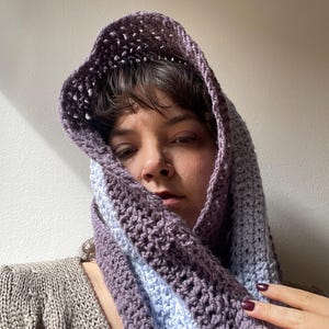 May include: A handmade crochet hood in shades of purple and light blue. The hood is draped over the head and shoulders. The crochet stitch creates a textured pattern. The hood is part of a scarf or cowl.