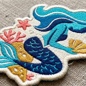 May include: Embroidered patch featuring a mermaid design. The mermaid has blue hair and a teal tail, with coral and starfish accents. The patch is outlined in white and has a nautical theme.