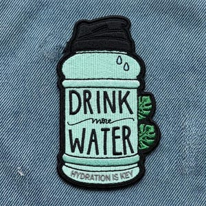 May include: Embroidered patch in the shape of a water bottle, with the words "DRINK more WATER" in black lettering. The patch is light blue with a black top and green leaf accents. The phrase "HYDRATION IS KEY" is at the bottom.