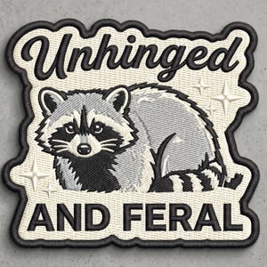 May include: An embroidered patch with a cream-colored background and a black border. It features a gray and black raccoon illustration, white star accents, and the black text "Unhinged" at the top and "AND FERAL" at the bottom in a bold font.