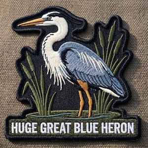 May include: Embroidered patch featuring a Great Blue Heron standing in a marsh. The bird has blue and white feathers, a long yellow beak, and orange legs. The text "HUGE GREAT BLUE HERON" is embroidered below the bird.