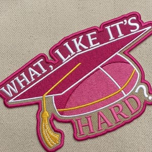 May include: Embroidered patch featuring a graduation cap design in shades of pink and white, with the text "WHAT, LIKE IT'S HARD?" in white lettering. The tassel is gold. The patch is outlined in a light brown color.