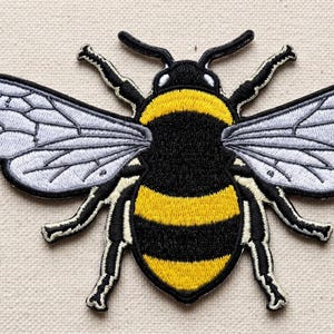 May include: Embroidered bee patch featuring a black body with yellow stripes, detailed wings, and black legs. The patch is designed with a white background and black outline, suitable for clothing or accessories.