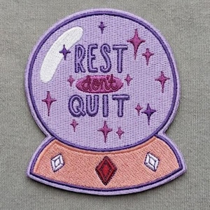 May include: Embroidered patch featuring a purple crystal ball design with the words "REST don't QUIT" in white and purple. The patch has a light purple background with star accents and a peach-colored base.