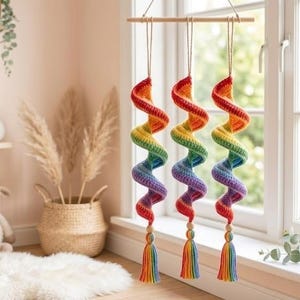 Op de afbeelding: Drie hangende spiraaldecoraties van gehaakt garen in een regenboogkleurverloop. Elke spiraal hangt aan een horizontale houten staaf met een dun koord en is voorzien van een houten kraal en een veelkleurige kwast aan de onderkant.