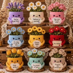 以下が含まれることがあります： 笑顔の刺繍が施された9つの編みぐるみの植木鉢。それぞれに花束とポジティブなメッセージが書かれた看板が付いています。鉢の色は紫、クリーム、ピンク、水色、黄色、赤、ゴールド、ミントグリーン、ベージュで、木製の棚に並べられています。