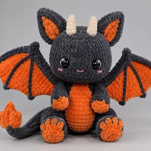 May include: A handmade crochet dragon plush toy featuring dark charcoal gray yarn with vibrant orange accents on the wings, belly, feet, and tail tip. The creature has two small cream-colored horns, large black glossy eyes, and pink stitched cheeks.