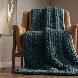 May include: A teal-colored, chunky knit blanket draped over a brown leather armchair. The blanket's texture is prominent, with thick, woven stitches. A small wooden side table with a mug and a painting are in the background.