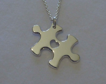 Puede incluir: Un collar de plata con un colgante de pieza de rompecabezas con un corazón recortado. El collar se muestra en una caja blanca con una cinta morada.
