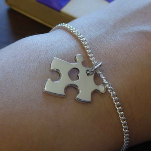 May include: A silver chain bracelet with a silver puzzle piece charm. The puzzle piece has a heart cut out of the centre. The bracelet is on a person's wrist.