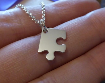 Puede incluir: Un collar de plata con un colgante de pieza de rompecabezas. El collar está en una mano.