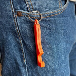 Könnte beinhalten: Ein leuchtend orangefarbener Flaschenöffner-Schlüsselanhänger hängt an einem silbernen Schlüsselring. Der Öffner besteht aus einem festen Material und hat ein einzigartiges, eckiges Design. Der Schlüsselanhänger ist an einer Jeans befestigt.
