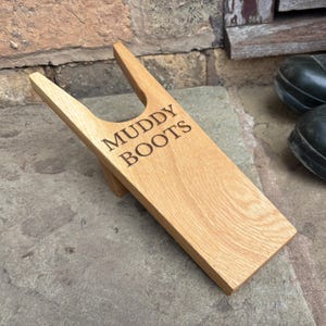 Könnte beinhalten: Ein hellbrauner Holz-Stiefelknecht mit der Gravur "MUDDY BOOTS". Der Stiefelknecht hat oben eine V-förmige Kerbe zum einfachen Herausziehen von Stiefeln. Er ist zum Entfernen von schmutzigen Stiefeln gedacht.