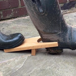 Könnte beinhalten: Ein hölzerner Stiefelknecht mit zwei dunkelgrünen Gummistiefeln. Der Stiefelknecht ist aus hellem Holz gefertigt und hat einen Schlitz, um die Ferse eines Stiefels zu halten. Ein Stiefel befindet sich auf dem Stiefelknecht, der andere daneben.