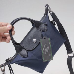 May include: Navy blue handbag with black leather accents, a top handle, and a detachable shoulder strap. The bag features a flap closure with a silver button and a tag that reads "ENERGY". The bag is made of a durable, textured material.