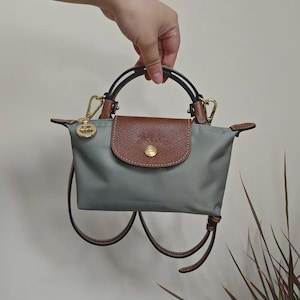 May include: A gray handbag with brown leather accents. The bag features a brown flap with a gold button closure and a gold charm. It has a top handle and a detachable shoulder strap.