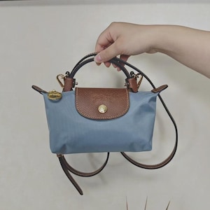 May include: A small, light blue handbag with brown leather accents. The bag features a brown flap with a gold-colored button closure and a matching handle. A detachable shoulder strap is also included.