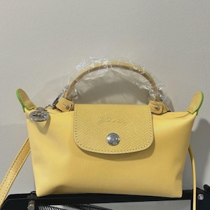 May include: A yellow handbag with a flap closure and a top handle. The bag has a detachable shoulder strap and a silver-tone metal clasp. The edges of the bag are accented with green trim. The brand name is embossed on the flap.